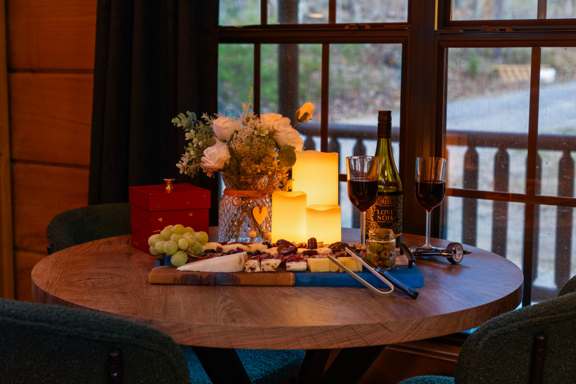 A candlelit charcuterie and wine table set beside a window at a Sky Mountain Retreat cabin