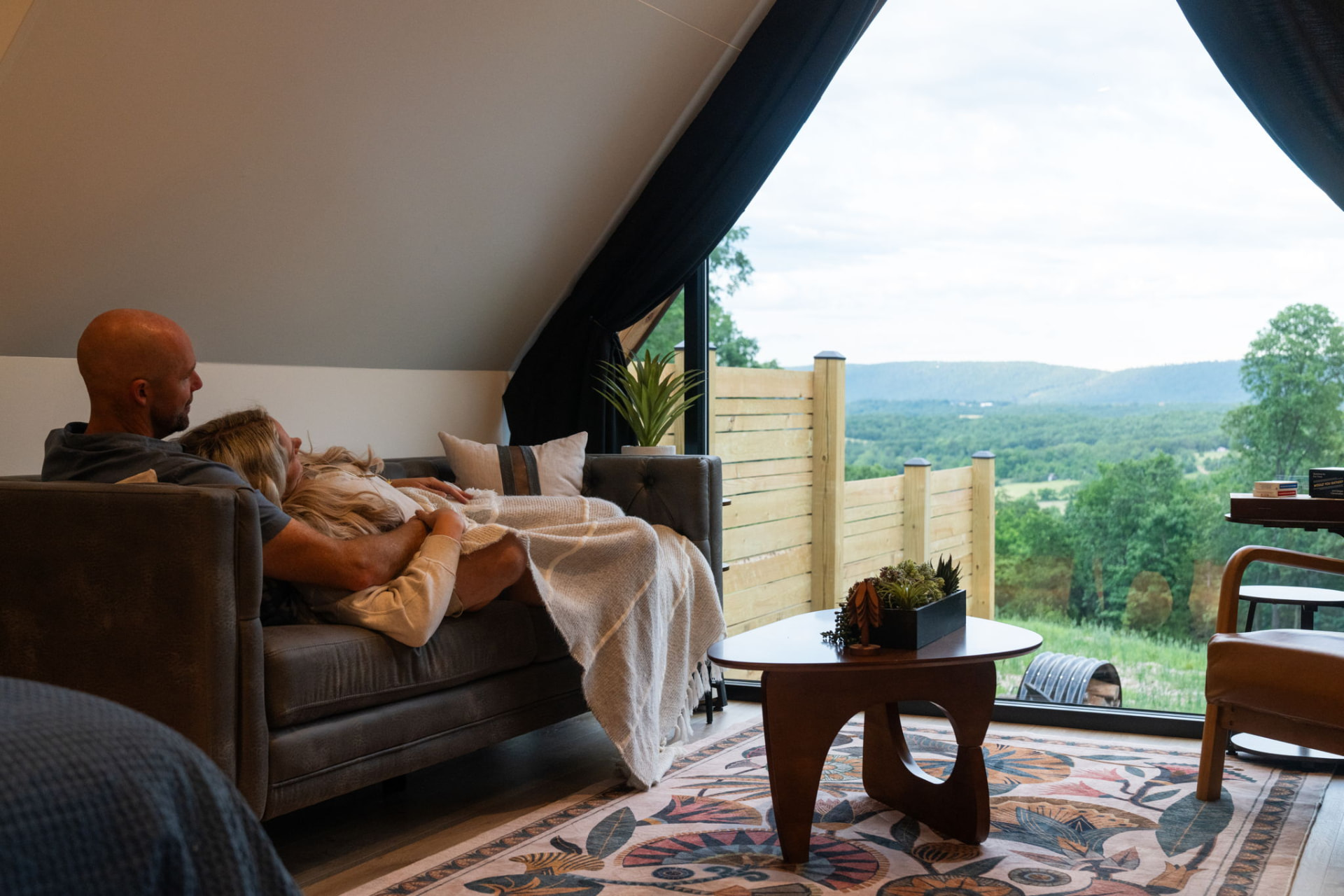 A couple wrapped in a blanket on the sofa at a Sky Mountain Retreat A-frame cabin, with the mountain view through the window