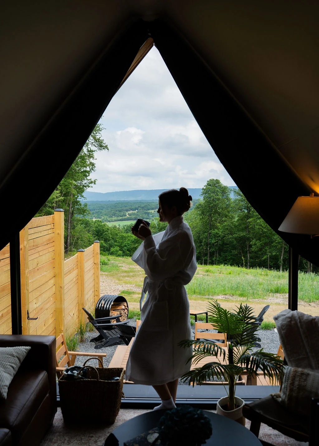 A guest in a white robe with a warm mug, looking out from an A-Frame toward the mountains
