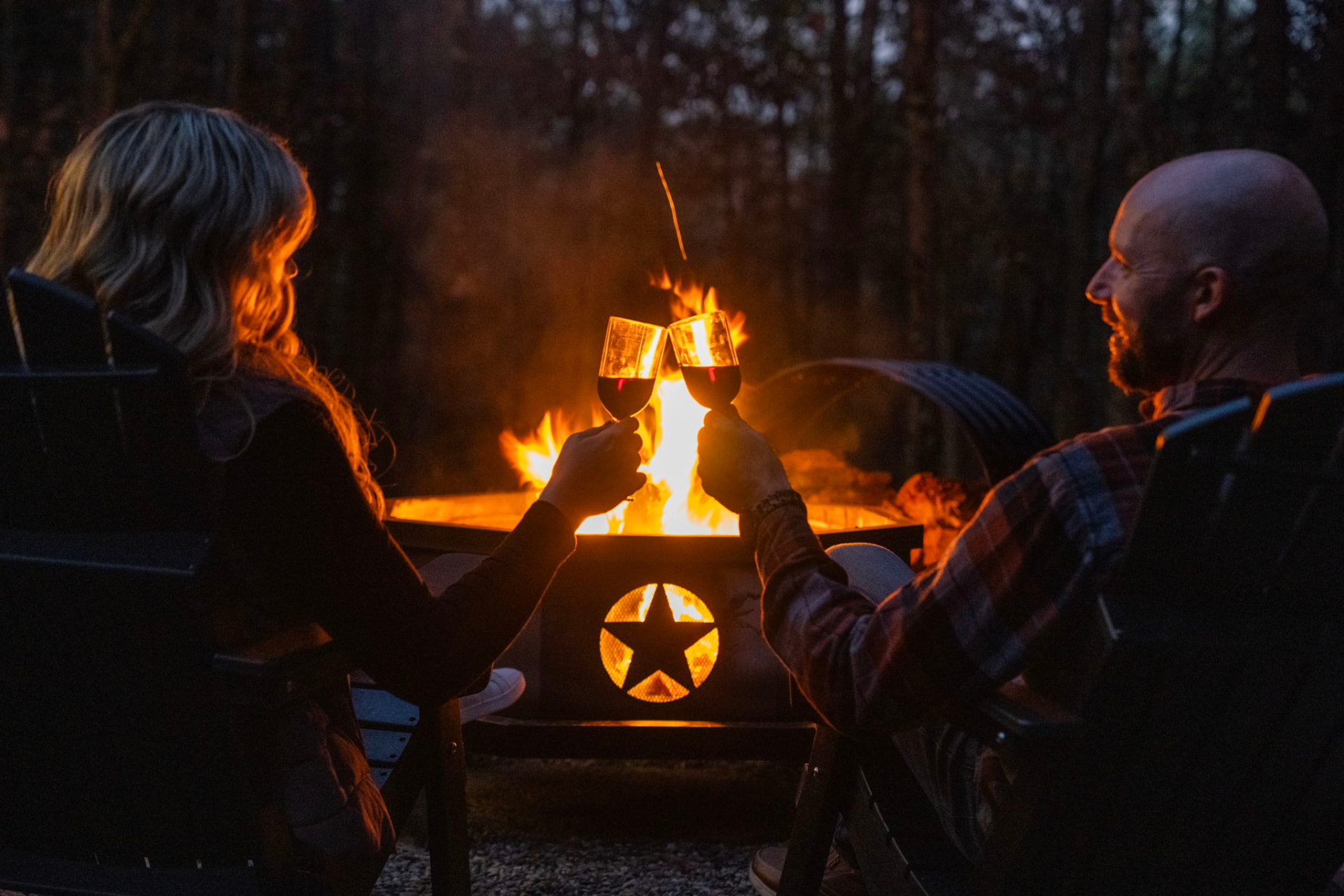 A couple toasting glasses of red wine beside a glowing fire pit at dusk